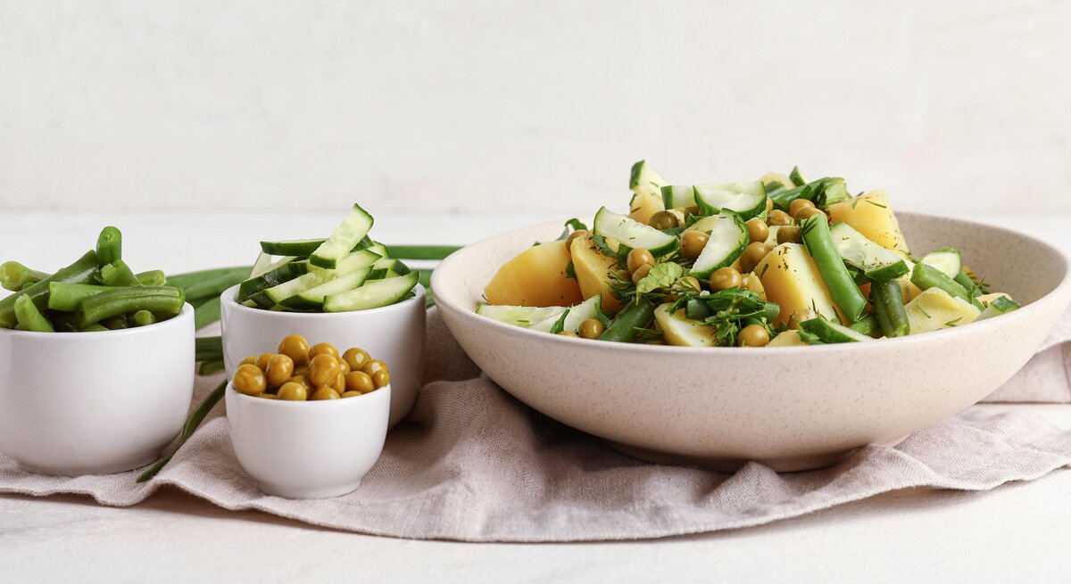 Eine weiße Schale mit dem Kartoffelsalat. Daneben drei kleine Schalen mit den weiteren Zutaten. Alles auf hellem Unter- und Hintergrund.