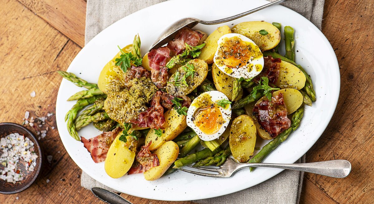 Kartoffelsalat mit gekochten Eiern, Pesto und knusprigem Pancetta und grünem Spargel auf einem weißen Teller mit Besteck. Daneben eine Schale mit Salz und mit Pesto. 