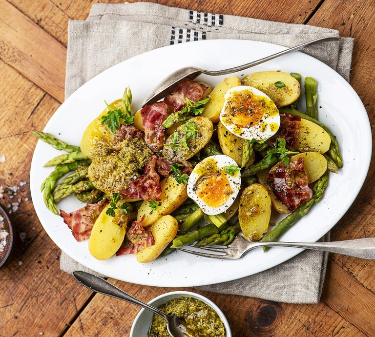 Kartoffelsalat mit gekochten Eiern, Pesto und knusprigem Pancetta und grünem Spargel auf einem weißen Teller mit Besteck. Daneben eine Schale mit Salz und mit Pesto.  | © StockFood / Schall, Ewgenija