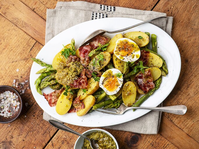 Kartoffelsalat mit gekochten Eiern, Pesto und knusprigem Pancetta und grünem Spargel auf einem weißen Teller mit Besteck. Daneben eine Schale mit Salz und mit Pesto.  | © StockFood / Schall, Ewgenija