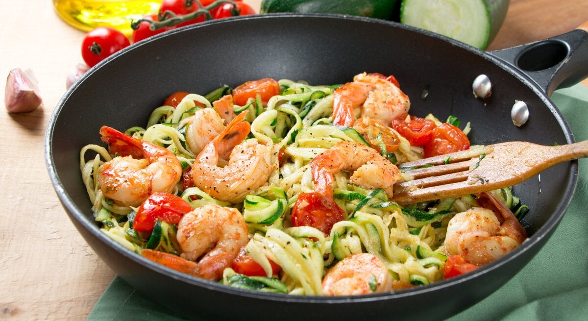 Auf einem Tisch steht eine Wok-Pfanne. darin sind Zoodles, Nudeln aus Zucchini, zu sehen. Auf dem Tisch liegen zwei Zucchini und Tomaten.