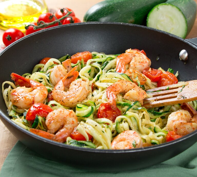 Auf einem Tisch steht eine Wok-Pfanne. darin sind Zoodles, Nudeln aus Zucchini, zu sehen. Auf dem Tisch liegen zwei Zucchini und Tomaten. | © Shutterstock/asife