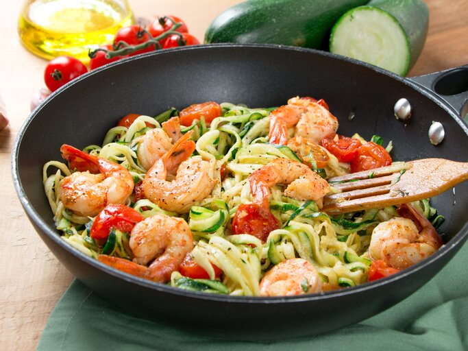 Auf einem Tisch steht eine Wok-Pfanne. darin sind Zoodles, Nudeln aus Zucchini, zu sehen. Auf dem Tisch liegen zwei Zucchini und Tomaten. | © Shutterstock/asife