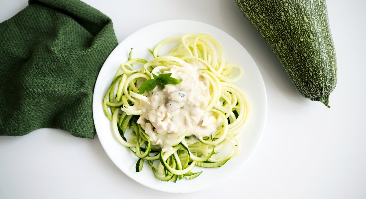 Auf einem weißen Untergrund liegen ein grünes Tuch und eine Zucchini. Daneben steht ein weißer Teller mit Zucchini-Nudeln mit Ziegenkäse-Soße.