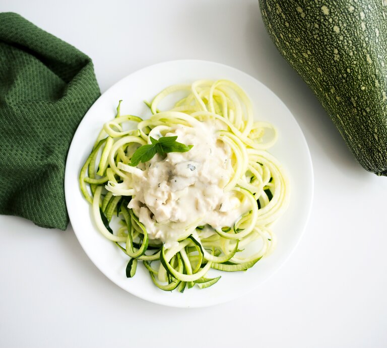 Auf einem weißen Untergrund liegen ein grünes Tuch und eine Zucchini. Daneben steht ein weißer Teller mit Zucchini-Nudeln mit Ziegenkäse-Soße. | © Shutterstock / Megan Betteridge