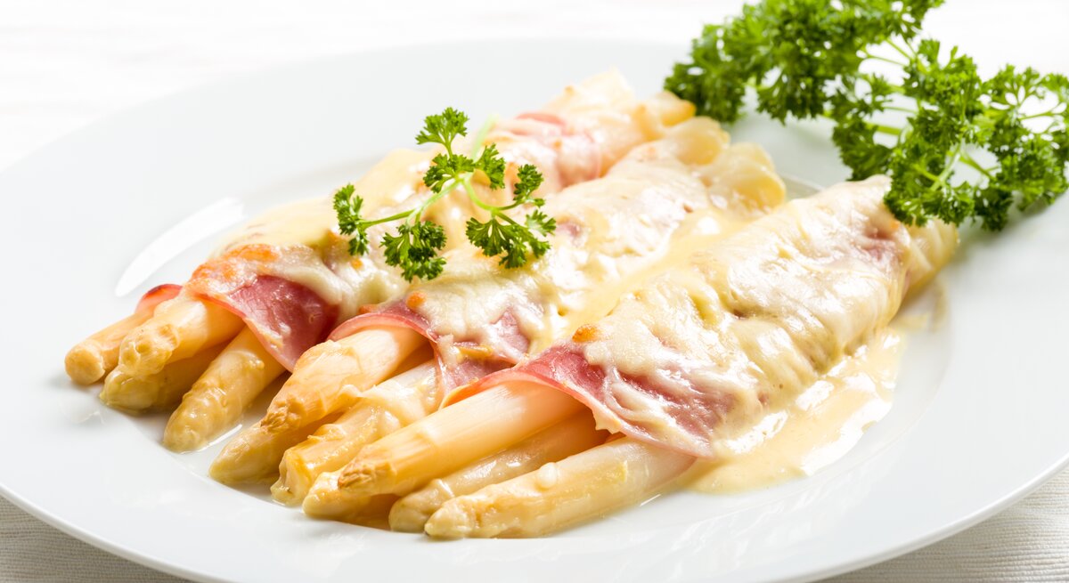 Weißer Spargel überbacken mit Schinken und Sauce hollandaise. Daneben liegt frische Petersilie.