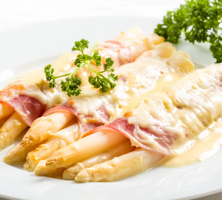 Weißer Spargel überbacken mit Schinken und Sauce hollandaise. Daneben liegt frische Petersilie. | © Shutterstock/Fischer Food Design