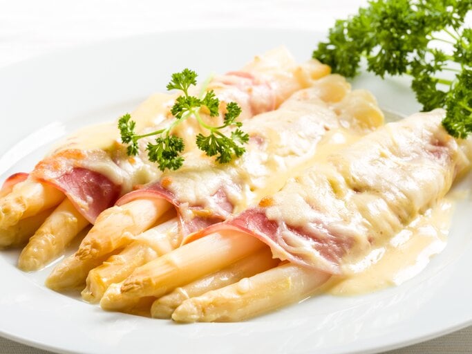 Weißer Spargel überbacken mit Schinken und Sauce hollandaise. Daneben liegt frische Petersilie. | © Shutterstock/Fischer Food Design