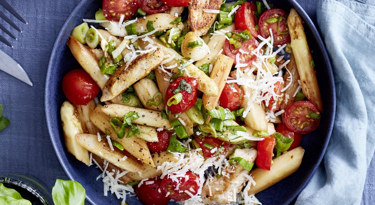 Italienischer Spargel-Mix mit Parmesan in dunkler Schüssel auf dunkelblauem Untergrund. Daneben Küchentuch, Besteck und Basilikumpflanze. 