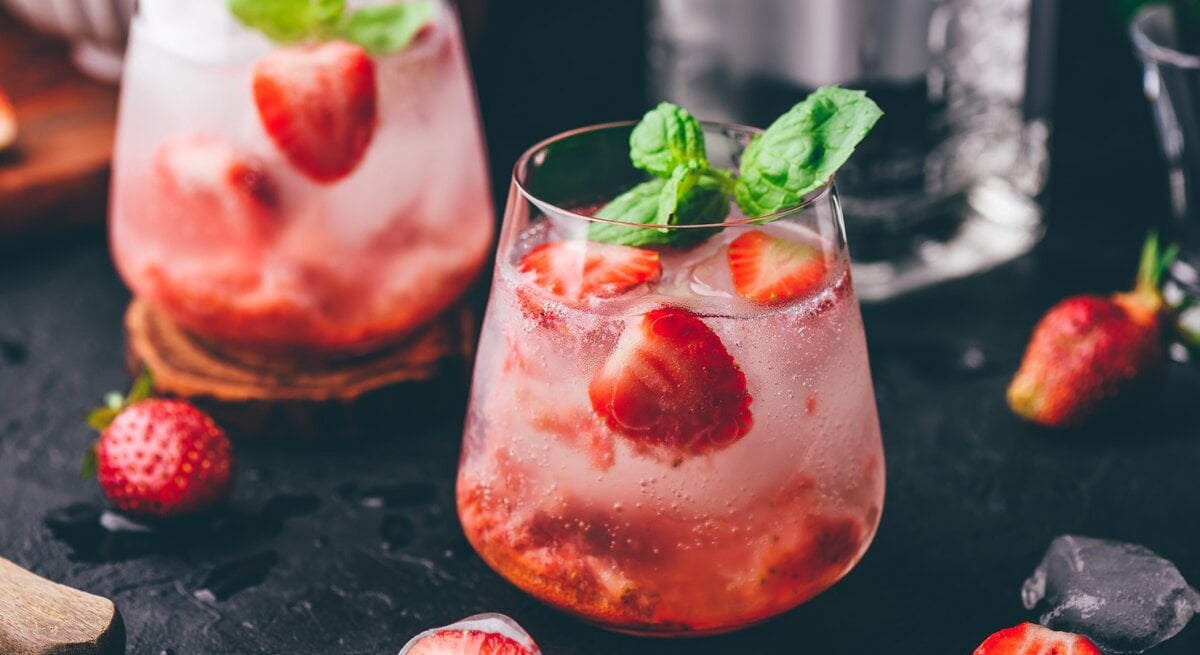 Erdbeer-Gin-Tonic auf einem dunklen Untergrund. Daneben liegen verschiedene Zutaten.