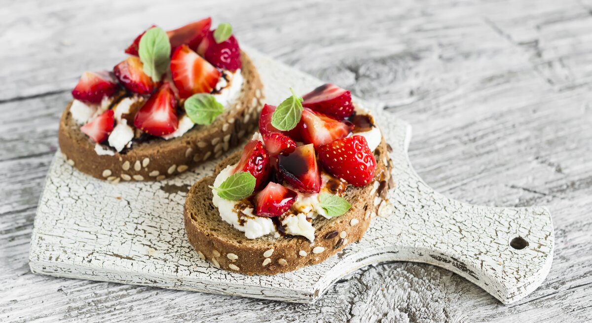 Draufsicht: zwei Erdbeer-Sandwiches mit Ricotta und Minze liegen auf einem hellen Holzbrett, das auf einem hellem Untergrund aus Holz liegt.