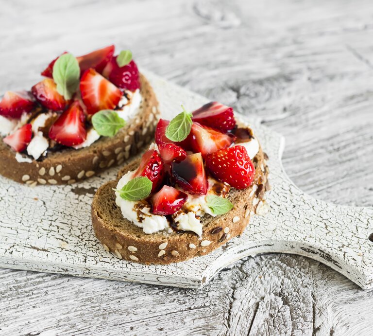 Draufsicht: zwei Erdbeer-Sandwiches mit Ricotta und Minze liegen auf einem hellen Holzbrett, das auf einem hellem Untergrund aus Holz liegt. | © Shutterstock / Kiian Oksana