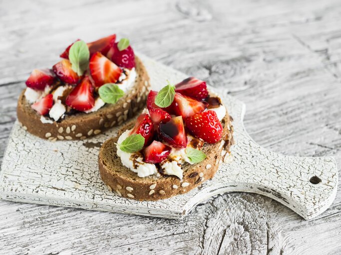 Draufsicht: zwei Erdbeer-Sandwiches mit Ricotta und Minze liegen auf einem hellen Holzbrett, das auf einem hellem Untergrund aus Holz liegt. | © Shutterstock / Kiian Oksana