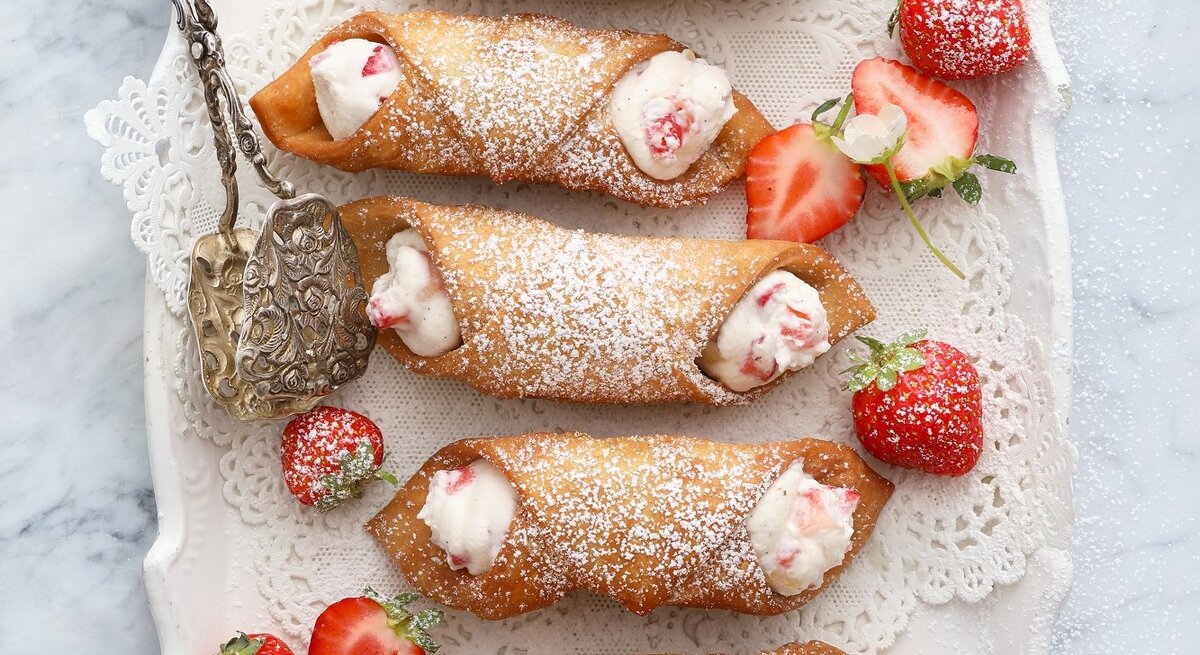 Italienische Cannoli mit Ricota-Erdbeer-Füllung, bestäubt mit Puderzucker und serviert auf einem weißen, verzierten Kuchenteller, angereicht mit frischen Erdbeeren. 