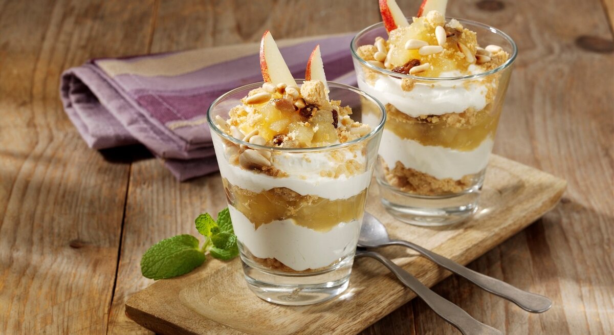 Auf einem Holzbrett stehen zwei Gläser, die mit Südtiroler Apfelstrudel aus dem Glas gefüllt sind. Sie sind mit Äpfeln dekoriert.