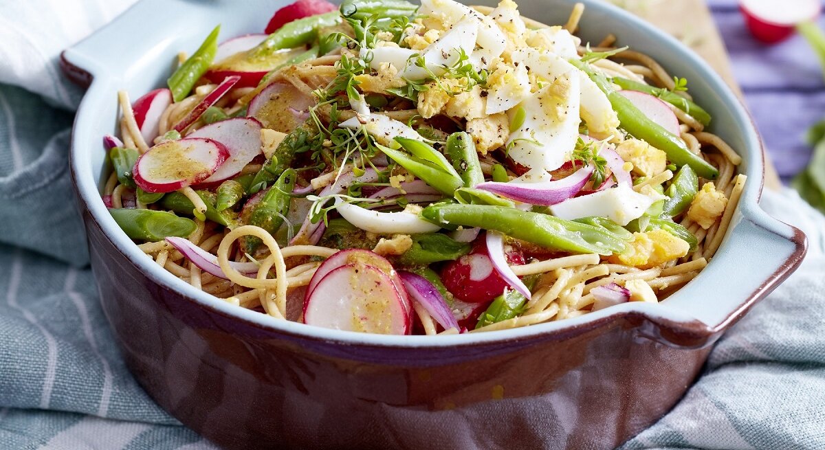 Eine Schüssel Bohnen-Nudel-Salat mit Eiern steht auf einem blau-weißem Tuch. Im Hintergrund sieht man Radieschen.