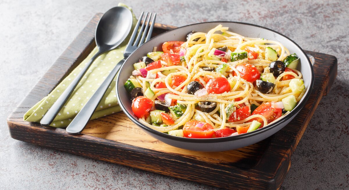 Kalifornischer Nudelsalat mit Zucchini, Paprika, Tomate, Zwiebel, Gurke und Oliven in einer Schale, angerichtet auf einem Holzbrett. Daneben liegt Besteck. 