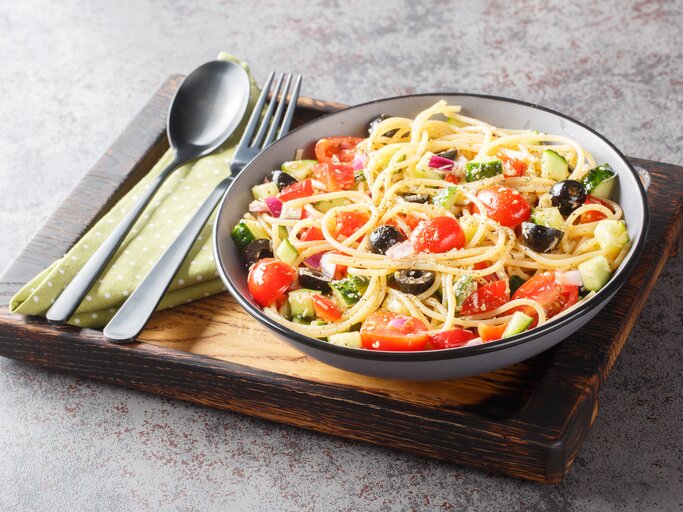 Kalifornischer Nudelsalat mit Zucchini, Paprika, Tomate, Zwiebel, Gurke und Oliven in einer Schale, angerichtet auf einem Holzbrett. Daneben liegt Besteck.  | © Getty Images/ALLEKO