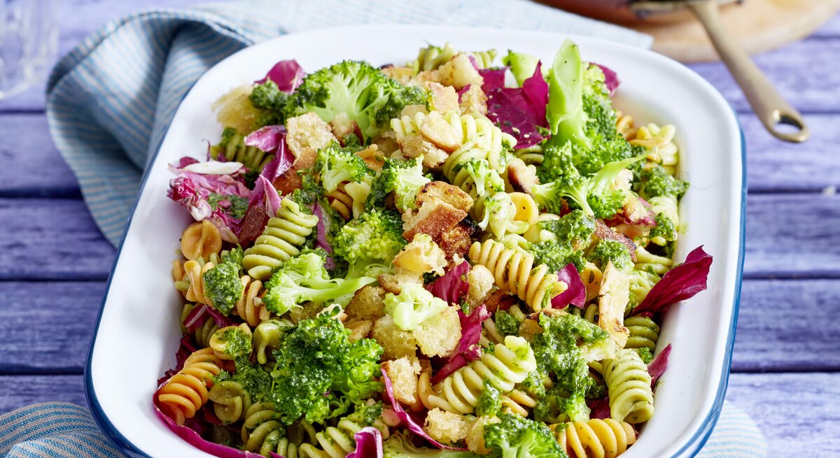 Eine weiße Auflaufform mit Brokkoli-Nudelsalat gefüllt auf einem violetten Tisch.