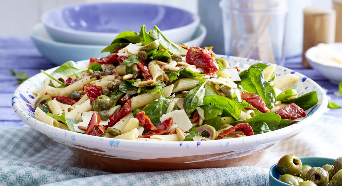 Italienischer Nudelsalat mit Kapern, Oliven, getrockneten Tomaten, Babyspinat und Parmesan in einer hellblauen Schale auf Küchentüchern gereicht. 