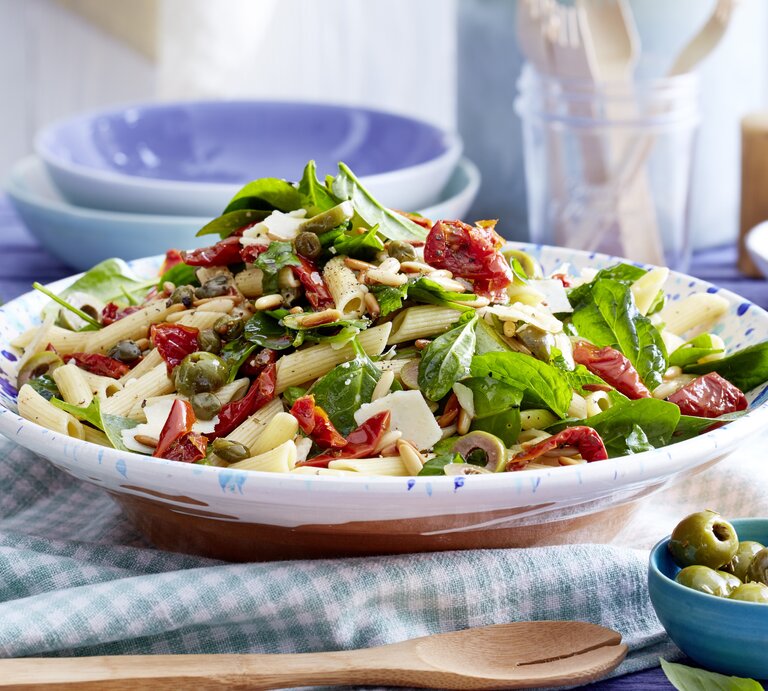 Italienischer Nudelsalat mit Kapern, Oliven, getrockneten Tomaten, Babyspinat und Parmesan in einer hellblauen Schale auf Küchentüchern gereicht.  | © EAT CLUB / Isabell Triemer