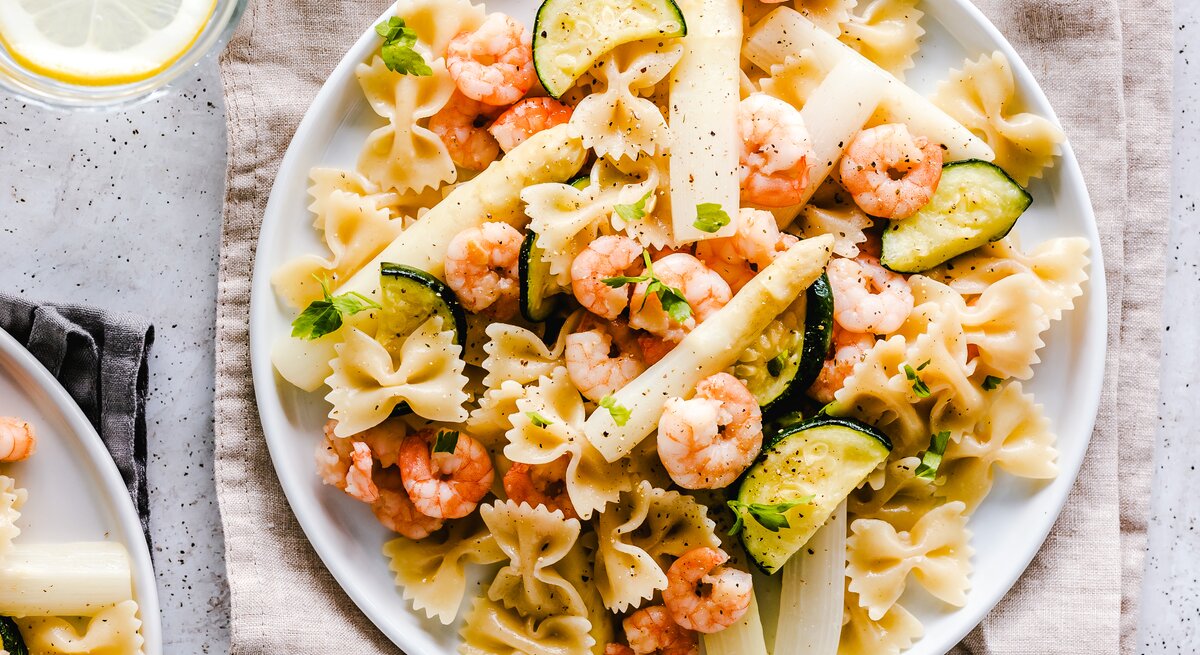 Teller mit Nudelsalat mit Spargel, Shrimps und Zucchini auf hellem Untergrund. Daneben steht ein Glas voll Wasser.
