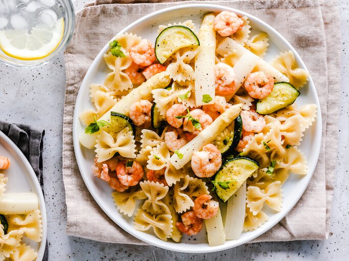Teller mit Nudelsalat mit Spargel, Shrimps und Zucchini auf hellem Untergrund. Daneben steht ein Glas voll Wasser. | © Getty Images/OatmealStories