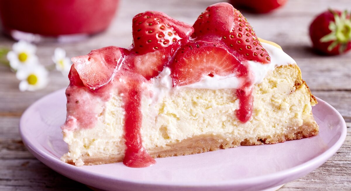 Auf einem Teller ist ein Stück Vanille-Erdbeer-Cheesecake serviert. Der Hintergrund ist hölzern und Gänseblümchen sind darauf verteilt. 