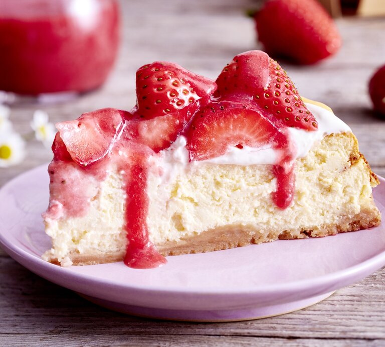 Auf einem Teller ist ein Stück Vanille-Erdbeer-Cheesecake serviert. Der Hintergrund ist hölzern und Gänseblümchen sind darauf verteilt.  | © EAT CLUB / Isabell Triemer