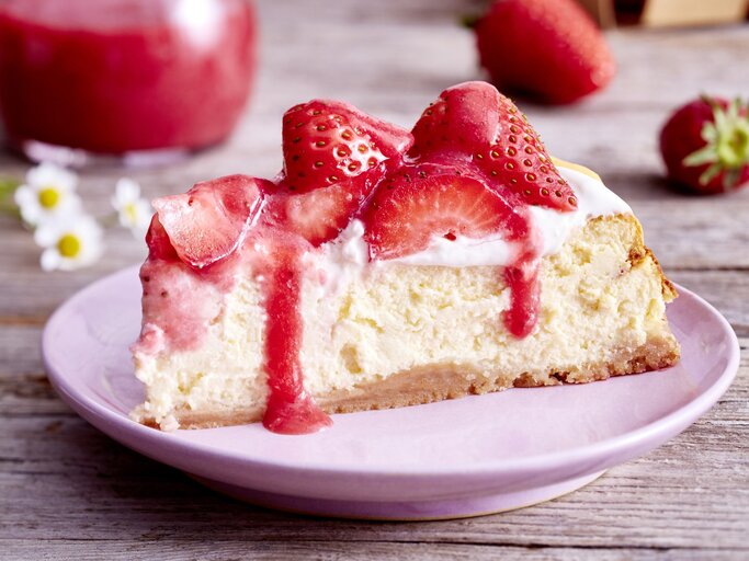 Auf einem Teller ist ein Stück Vanille-Erdbeer-Cheesecake serviert. Der Hintergrund ist hölzern und Gänseblümchen sind darauf verteilt.  | © EAT CLUB / Isabell Triemer