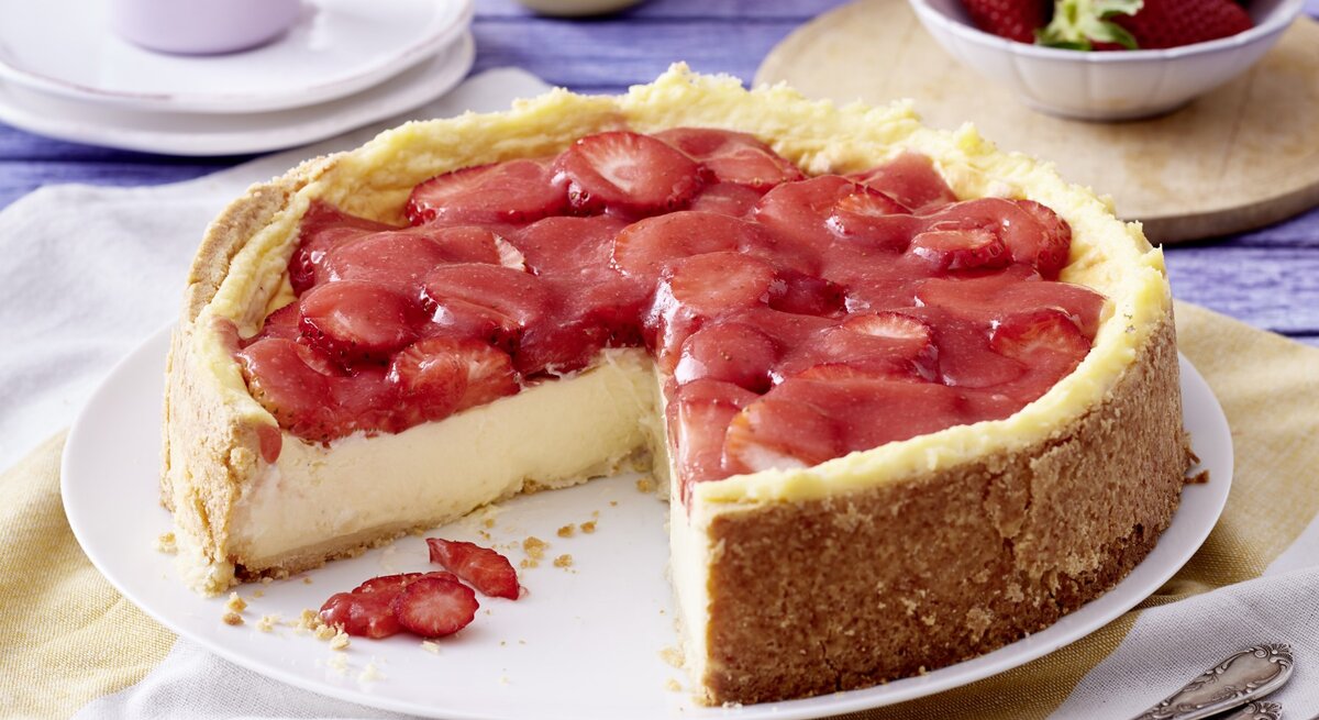 Angeschnittener Erdbeer-Schmandkuchen auf weißem Teller, daneben Gabeln, Geschirr und Erdbeeren auf Küchentüchern mit blauem Holzuntergrund. 