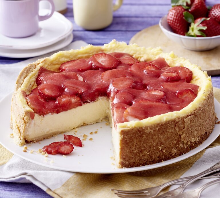 Angeschnittener Erdbeer-Schmandkuchen auf weißem Teller, daneben Gabeln, Geschirr und Erdbeeren auf Küchentüchern mit blauem Holzuntergrund.  | © EAT CLUB / Isabell Triemer