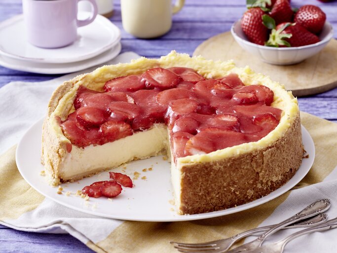 Angeschnittener Erdbeer-Schmandkuchen auf weißem Teller, daneben Gabeln, Geschirr und Erdbeeren auf Küchentüchern mit blauem Holzuntergrund.  | © EAT CLUB / Isabell Triemer