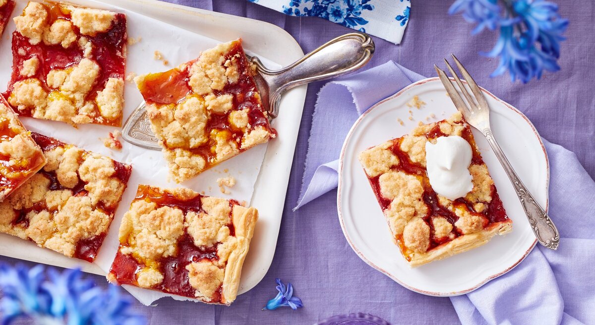 Ein weißes Kuchenblech mit Stücken des Prasselkuchens mit Erbeeren und Mango darauf neben einem Teller mit einem weiterem Stück, einer Gabel und blauen Blumen, vor einem fliederfarbenen Hintergrund, von oben. 