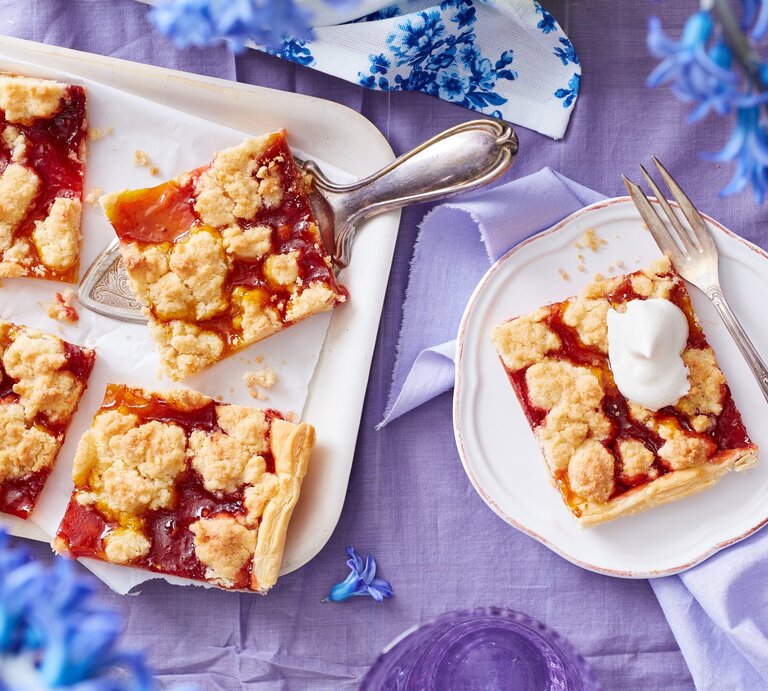 Ein weißes Kuchenblech mit Stücken des Prasselkuchens mit Erbeeren und Mango darauf neben einem Teller mit einem weiterem Stück, einer Gabel und blauen Blumen, vor einem fliederfarbenen Hintergrund, von oben.  | © EAT CLUB/Isabell Triemer