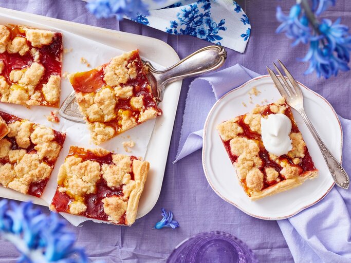 Ein weißes Kuchenblech mit Stücken des Prasselkuchens mit Erbeeren und Mango darauf neben einem Teller mit einem weiterem Stück, einer Gabel und blauen Blumen, vor einem fliederfarbenen Hintergrund, von oben.  | © EAT CLUB/Isabell Triemer