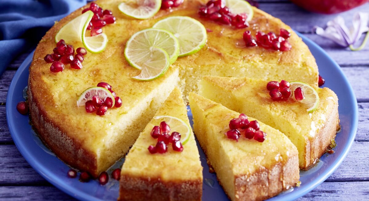Saftiger Joghurt-Limetten-Kuchen mit Granatapfelkernen und gezuckerten Limettenscheiben, angechnitten serviert auf einem blauenTeller. 