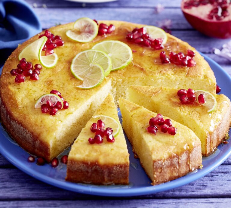 Saftiger Joghurt-Limetten-Kuchen mit Granatapfelkernen und gezuckerten Limettenscheiben, angechnitten serviert auf einem blauenTeller.  | © EAT CLUB / Isabell Triemer