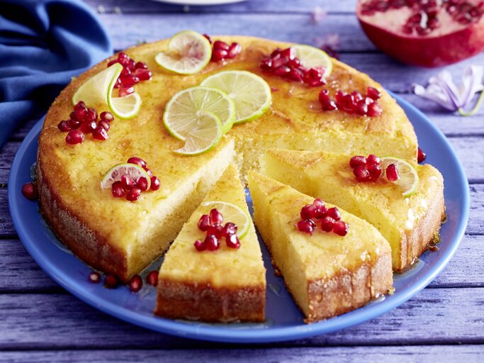 Saftiger Joghurt-Limetten-Kuchen mit Granatapfelkernen und gezuckerten Limettenscheiben, angechnitten serviert auf einem blauenTeller.  | © EAT CLUB / Isabell Triemer