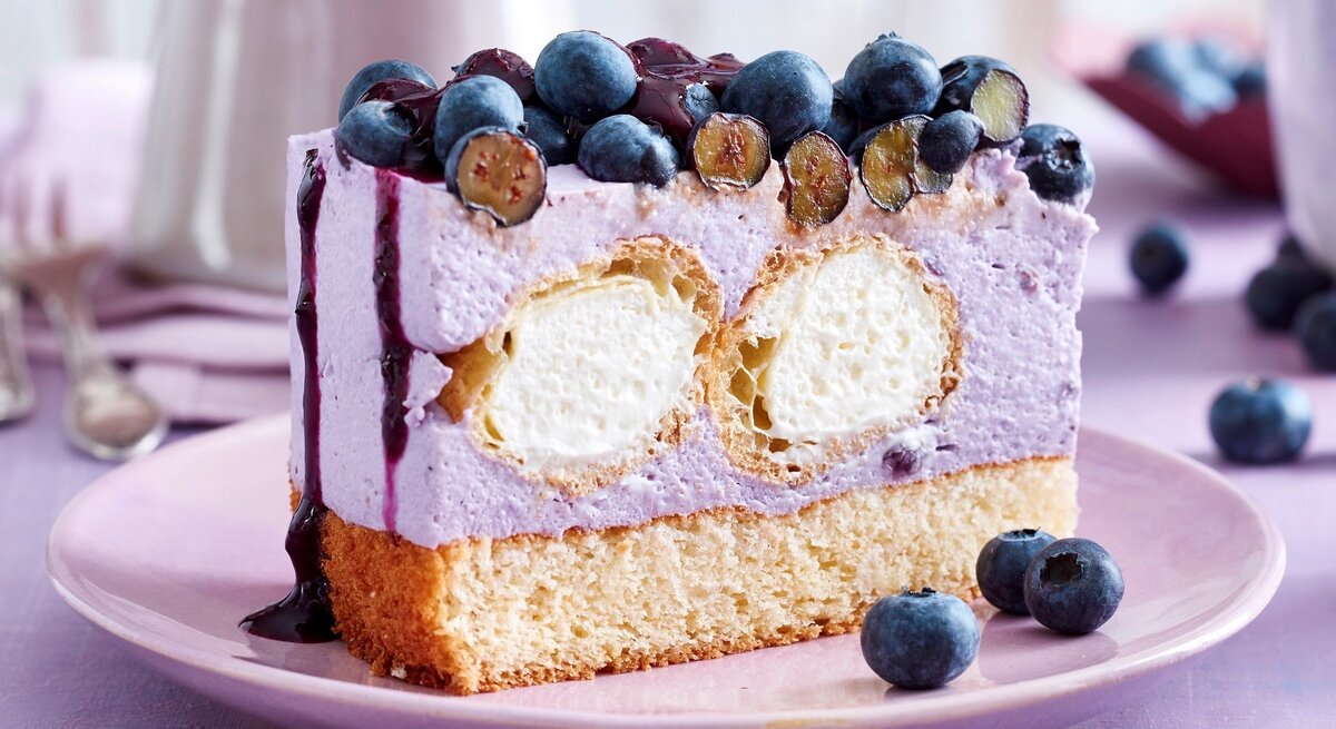 Auf einem Teller steht ein Stück Windbeutel-Joghurt-Torte. Es ist mit Blaubeeren und Soße verfeinert. Im Inneren befinden sich zwei Windbeutel.