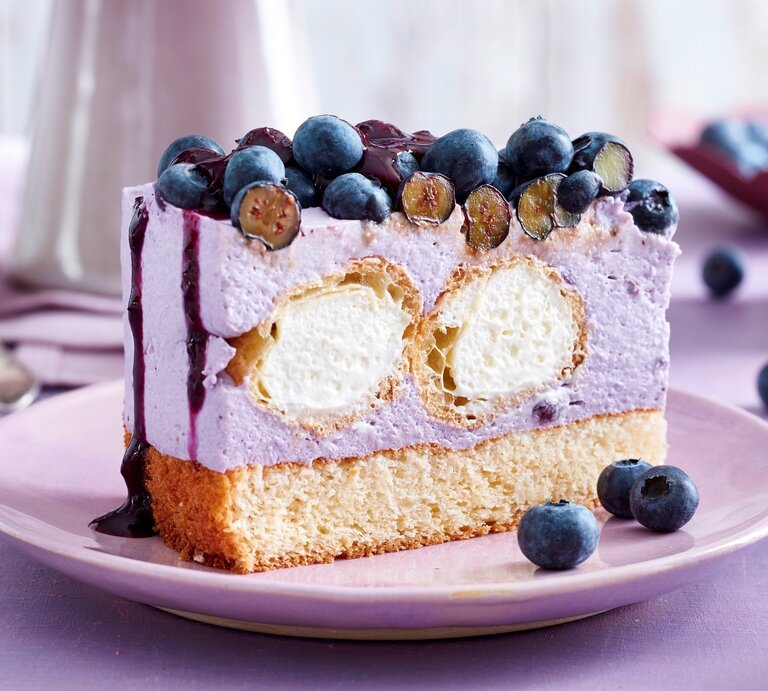 Auf einem Teller steht ein Stück Windbeutel-Joghurt-Torte. Es ist mit Blaubeeren und Soße verfeinert. Im Inneren befinden sich zwei Windbeutel. | © EAT CLUB / Isabell Triemer