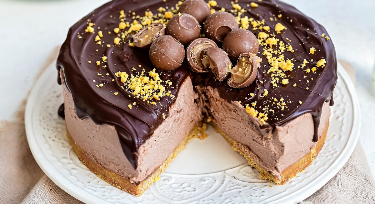 Ein weißer Teller mit einem angeschnittenen Baileys-Käsekuchen mit Schokoglasur auf einem braunen Tischtuch vor einem hellen Hintergrund. 
