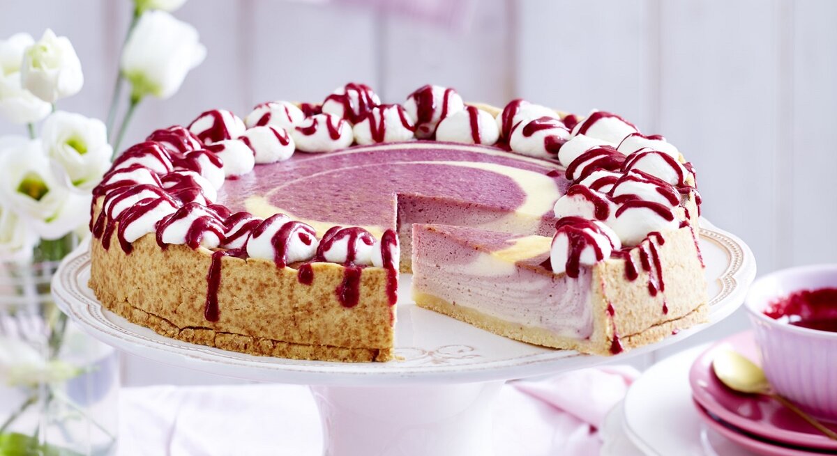 Auf einem Tortenteller steht ein beeriger Zebra-Käsekuchen. Er ist mit Sahnehäubchen und Soße verziert.