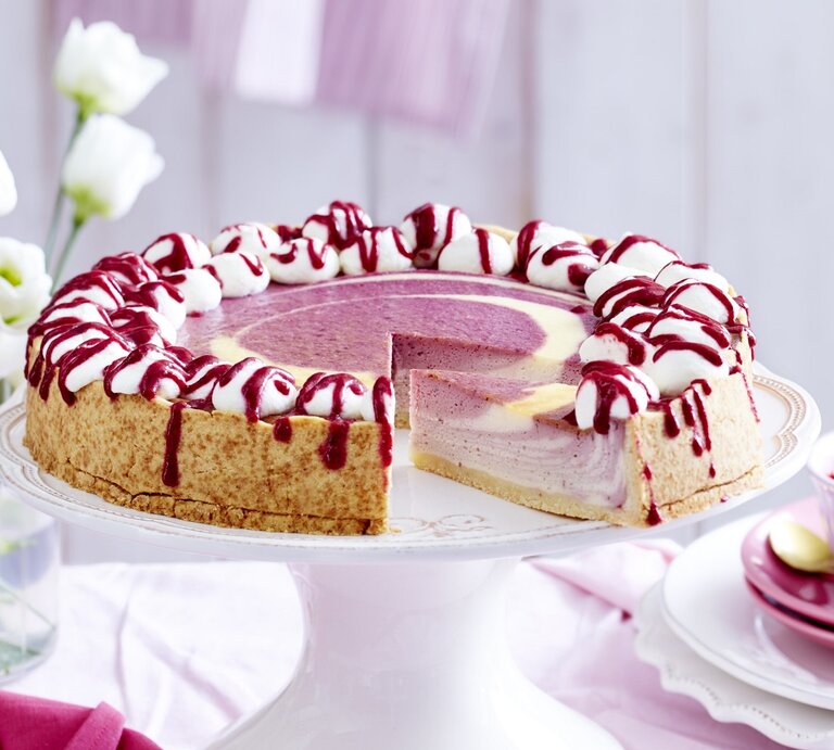 Auf einem Tortenteller steht ein beeriger Zebra-Käsekuchen. Er ist mit Sahnehäubchen und Soße verziert. | © EAT CLUB / Isabell Triemer