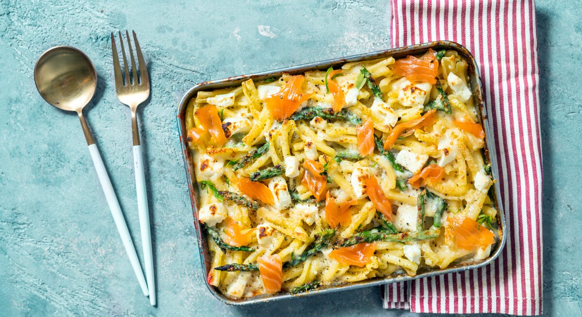 Spargel-Nudel-Auflauf mit Lachs in Auflaufform auf hellblauem Untergrund. Daneben liegen ein rot-weiß-gestreiftes Geschirrtuch sowie Löffel und Gabel. 