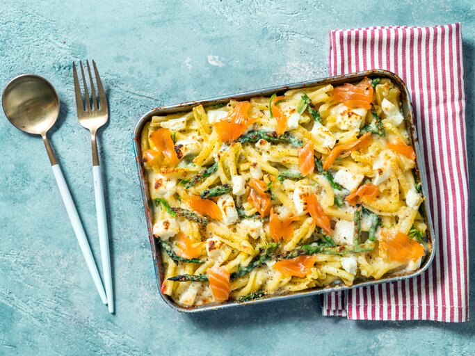 Spargel-Nudel-Auflauf mit Lachs in Auflaufform auf hellblauem Untergrund. Daneben liegen ein rot-weiß-gestreiftes Geschirrtuch sowie Löffel und Gabel.  | © Getty Images/Carlo A