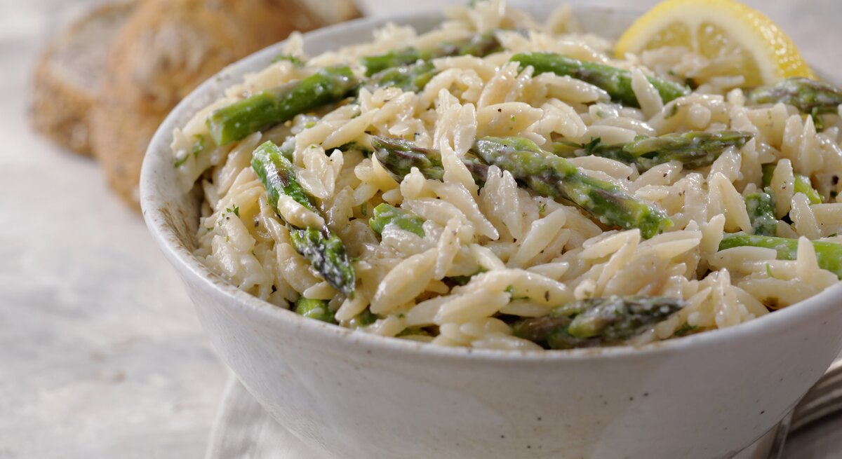 Im Vordergrund steht eine weiße Schüssel mit einem cremigen Spargel-Orzo-Topf mit Parmesan. Daneben liegen zwei Brötchen.