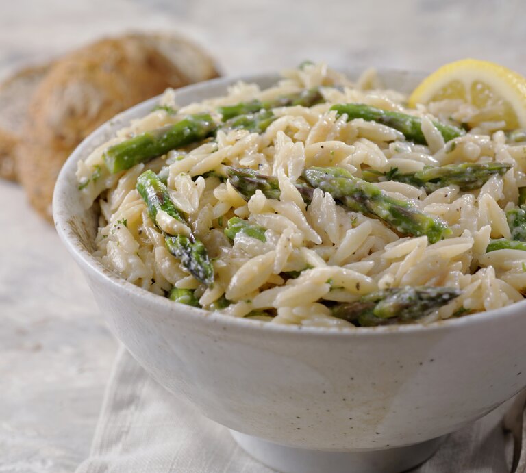 Im Vordergrund steht eine weiße Schüssel mit einem cremigen Spargel-Orzo-Topf mit Parmesan. Daneben liegen zwei Brötchen. | © Getty Images / LauriPatterson