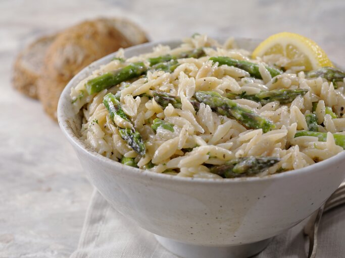 Im Vordergrund steht eine weiße Schüssel mit einem cremigen Spargel-Orzo-Topf mit Parmesan. Daneben liegen zwei Brötchen. | © Getty Images / LauriPatterson