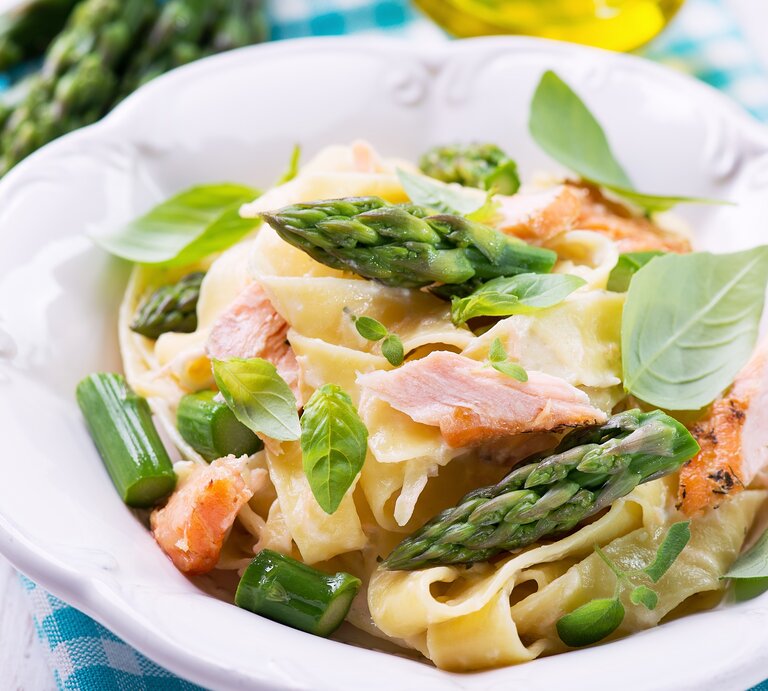 Ein weißer Teller mit dem Pastagericht auf einem hellen Holztisch mit blau-weiß-karierter Decke. Hinten unscharf Olivenöl und grüner Spargel zu erkennen. | © Shutterstock/ Kamila i Wojtek Cyganek
